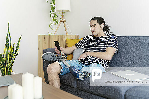 Young man with prosthetic leg using smart phone on sofa at home