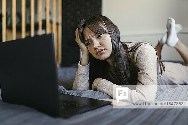 Young woman with head in hand using laptop on bed at home