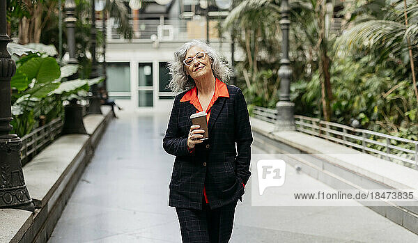 Confident mature businesswoman with disposable cup walking on footpath