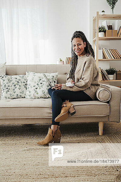 Lächelnde Frau mit Laptop sitzt zu Hause auf dem Sofa