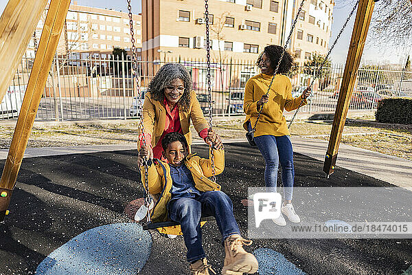Lächelnde Frau schaut Mutter zu  die ihren Enkel auf der Schaukel auf dem Spielplatz antreibt