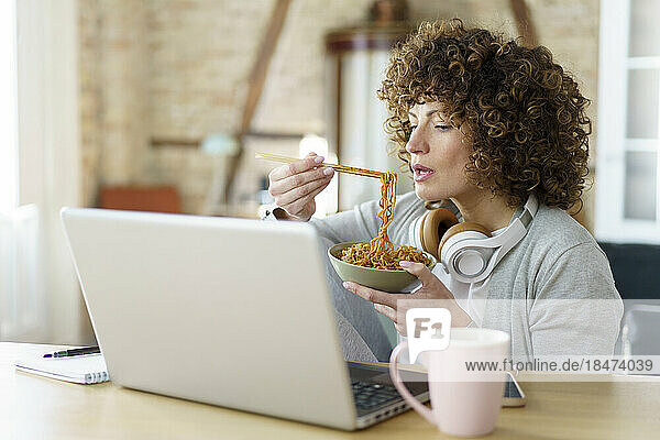 Freelancer with bowl of noodles working on laptop at home
