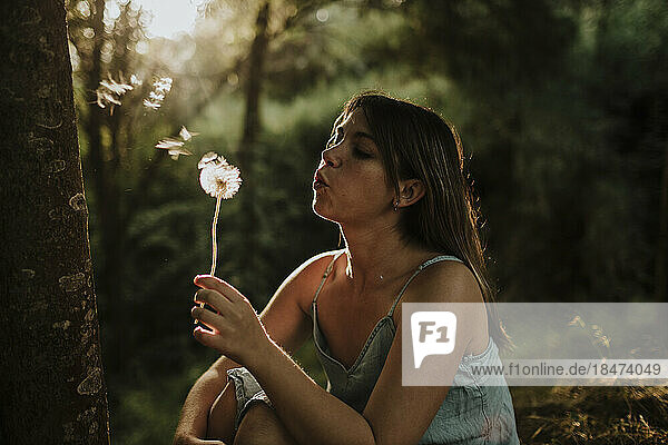 Young woman blowing on dandelion flower in forest