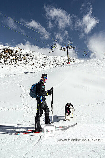 Lächelnder Mann steht mit Skistöcken und Hund im Schnee