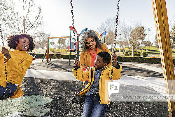 Smiling woman looking at grandmother pushing son on swing on playground