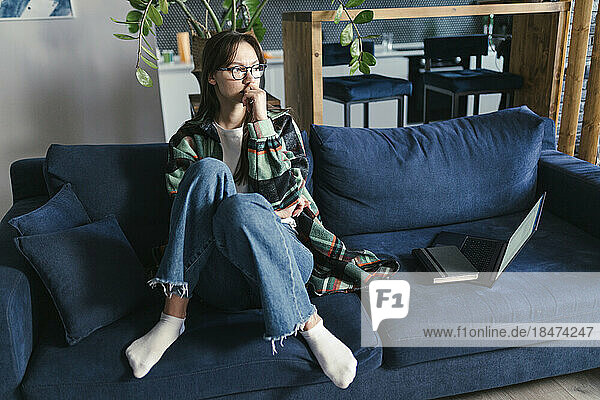 Thoughtful young woman sitting on sofa with laptop at home
