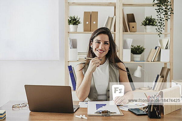 Glückliche Geschäftsfrau sitzt mit Klemmbrett am Schreibtisch im Büro