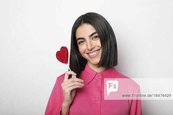 Smiling woman holding heart shaped lollipop against white background