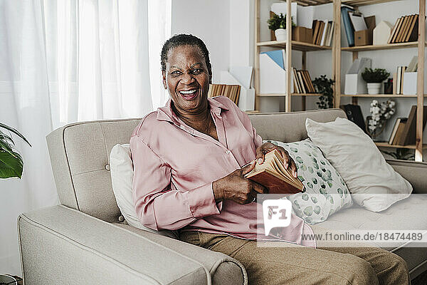 Glückliche ältere Frau mit Buch sitzt zu Hause auf dem Sofa