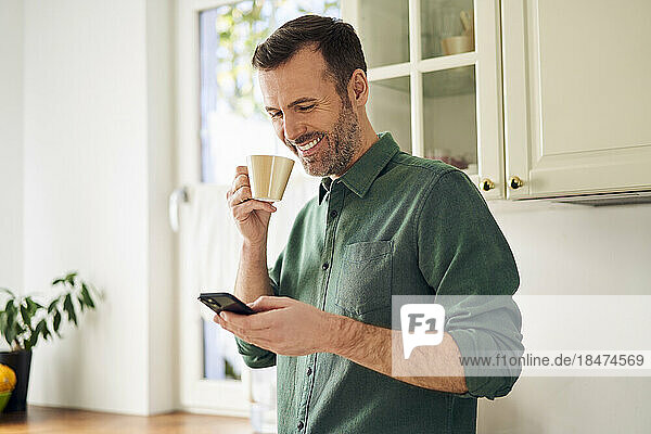 Smiling man using phone while drinking coffee in his kitchen