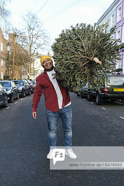 Happy man carrying Christmas tree standing on street