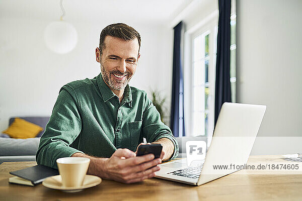 Selbstbewusster Mann sitzt zu Hause am Tisch und benutzt Laptop und bezahlt mit Mobiltelefon