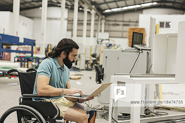 Engineer sitting in wheelchair examining machine through laptop in factory