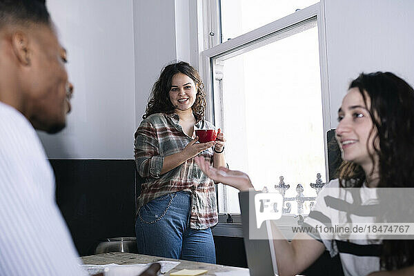 Lächelnde Geschäftsfrau mit Kaffeetasse und Blick auf Kollegen  die im Büro diskutieren