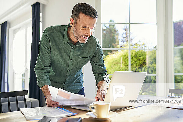 Smiling man doing paperwork at home using laptop