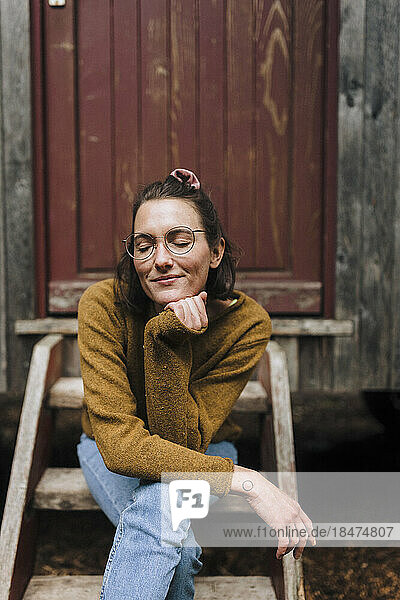 Smiling woman with eyes closed and hand on chin sitting in front of cabin