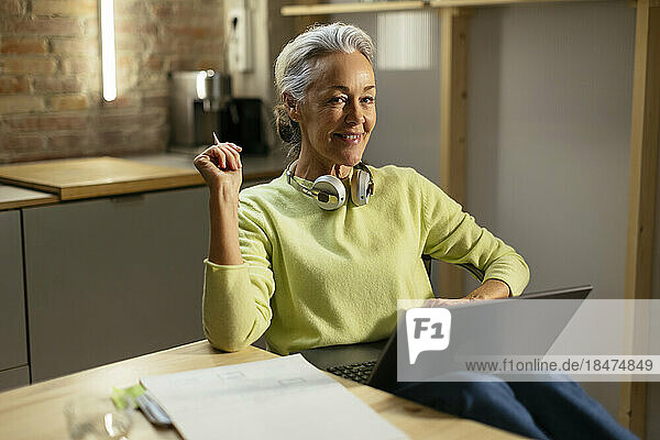 Smiling freelancer with laptop sitting at desk in kitchen
