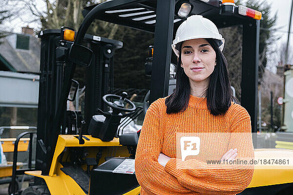 Confident young woman standing with arms crossed in front of forklift