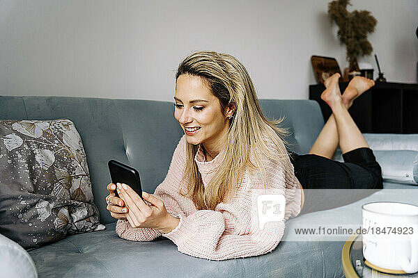 Smiling young woman using smart phone lying on sofa at home
