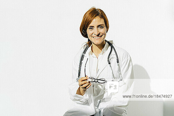 Happy female doctor with eyeglasses and stethoscope in front of white wall
