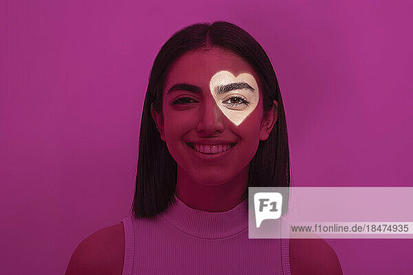 Lächelnde Frau mit herzförmigem Licht auf dem Auge vor rosa Hintergrund