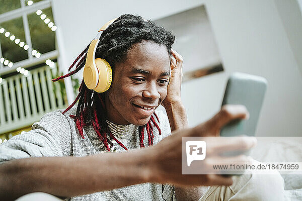 Smiling young man wearing headphones using smart phone at home