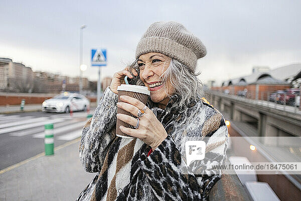 Happy mature woman with disposable cup talking through smart phone on footpath