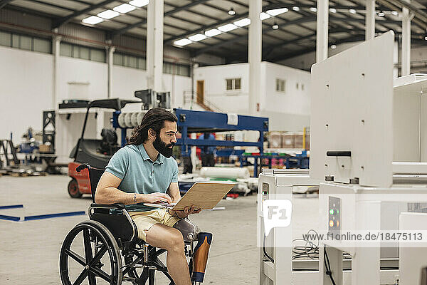 Engineer sitting in wheelchair analyzing machine through laptop in factory