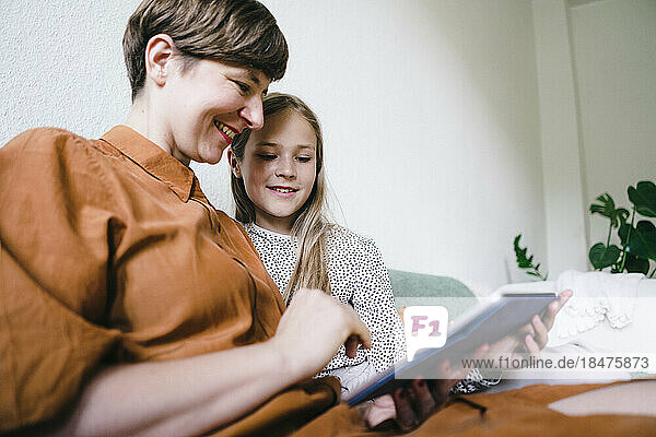 Lächelnde Frau teilt Tablet-PC mit Tochter  die zu Hause sitzt
