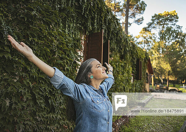 Smiling mature woman standing at overgrown facade with outstretched arms