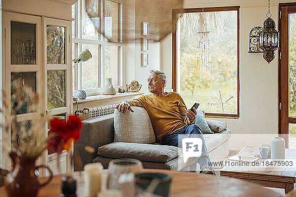 Thoughtful senior man sitting with smart phone on sofa at home