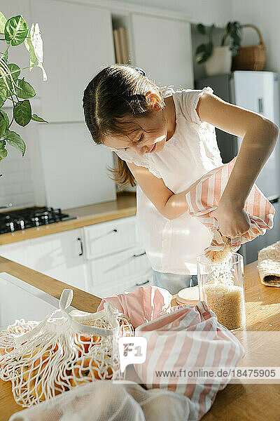Girl pouring rice from reusable bag in glass jar at home