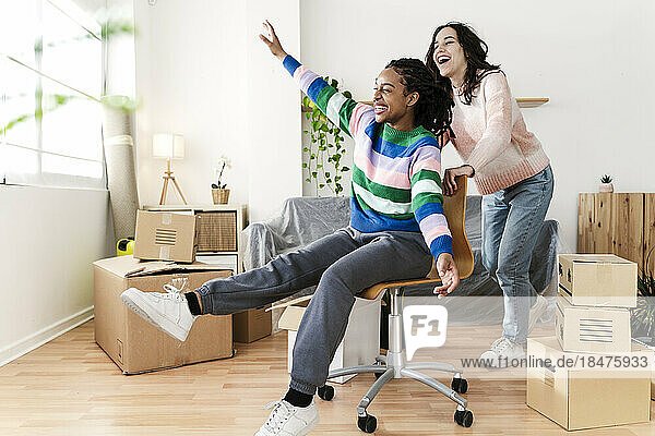 Carefree young woman pushing friend sitting in swivel chair at new home