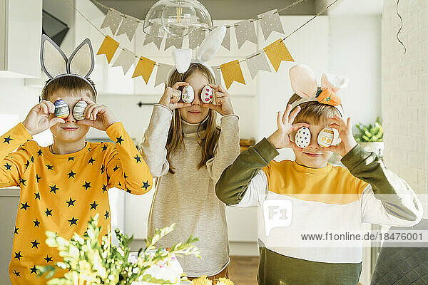 Smiling boys and girl holding Easter eggs over eyes at home