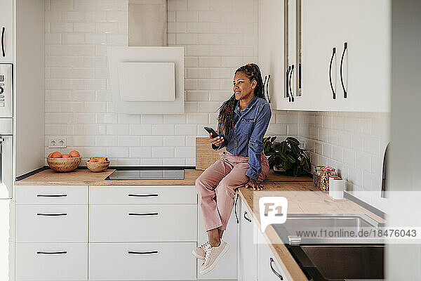 Smiling woman sitting on kitchen counter with smart phone at home