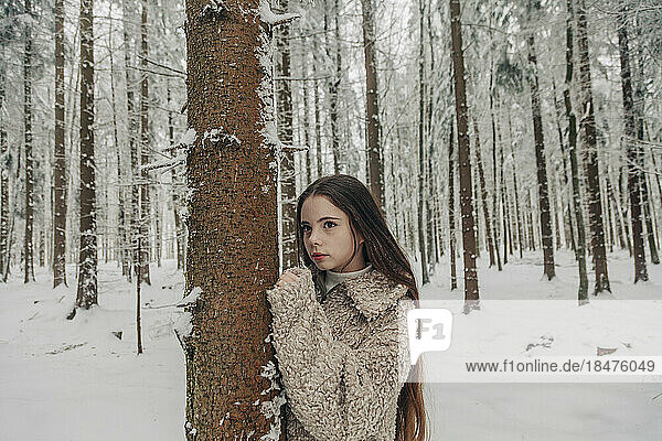 Contemplative teenage girl standing by tree in snowy forest