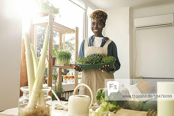 Smiling woman holding container of microgreens at home