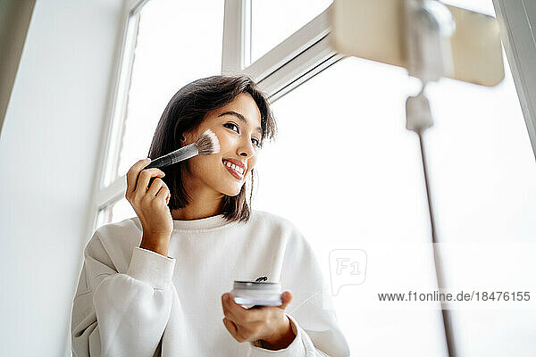 Young influencer applying blusher and filming through mobile phone at home