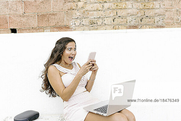 Surprised woman using smart phone sitting with laptop on bench