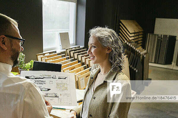 Smiling businesswoman looking at colleague holding construction plan in architect's office