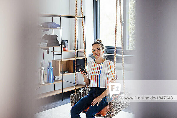 Lächelnde Geschäftsfrau mit Smartphone sitzt auf Schaukel im Büro