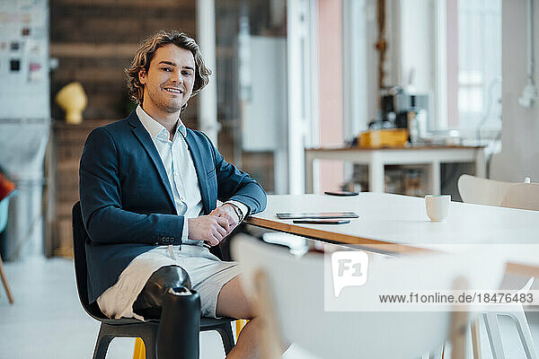 Lächelnder junger Geschäftsmann mit gefalteten Händen sitzt auf einem Stuhl im Büro