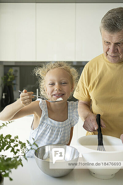Playful girl holding spoon by grandfather preparing batter in kitchen