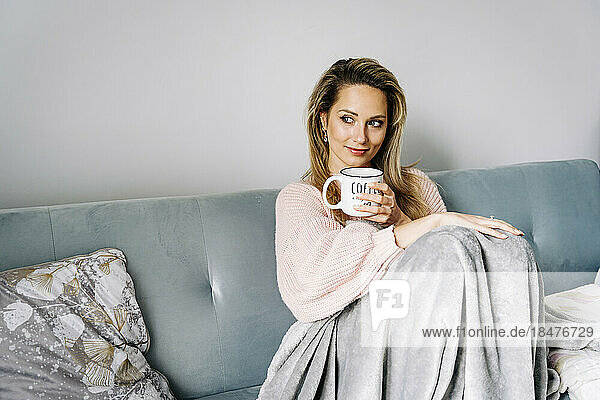 Thoughtful smiling woman sitting with coffee cup on sofa at home