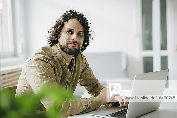 Smiling young businessman sitting with laptop at desk in office