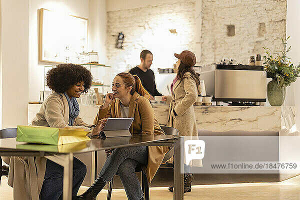 Happy young woman using tablet PC with friend sitting at table in cafe
