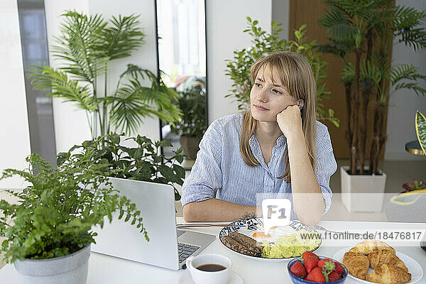 Contemplative freelancer sitting with breakfast and laptop at desk