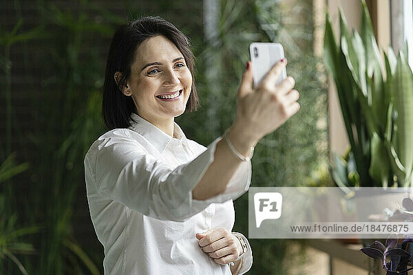 Happy woman taking selfie through smart phone in front of plants