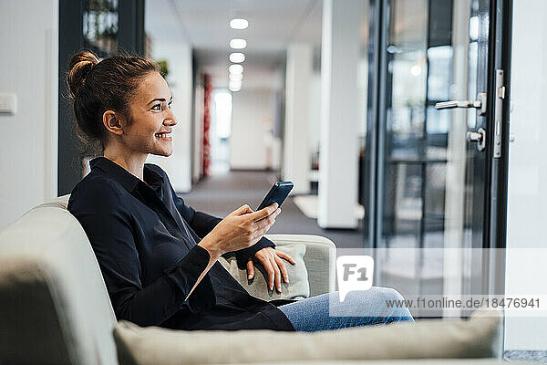 Smiling young businesswoman sitting with smart phone on couch in office