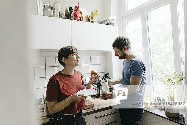 Woman eating food standing by man in kitchen